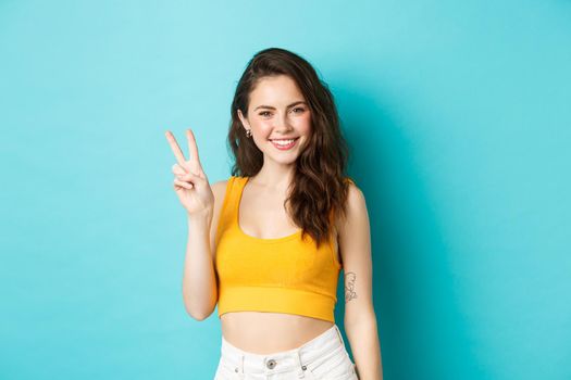 Summer and lifestyle concept. Beautiful girl tourist in crop top, posing with v-sign and smiling happy at camera, standing against blue background.