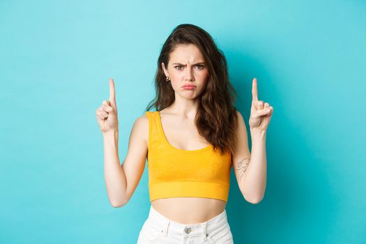 Summer lifestyle concept. Disappointed moody girl complaining about unfair situation, frowning and sulking upset, pointing fingers up at logo, standing over blue background.