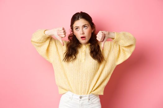 How lame. Displeased and indifferent young woman roll eyes, sighing and look aside, showing thumbs down with skeptical face, standing against pink background.