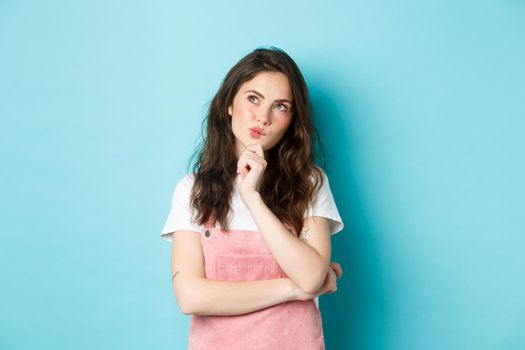 Thoughtful brunette woman making choice, holding hand on chin and thinking, looking at upper left corner with pensive face, consider something, standing over blue background.