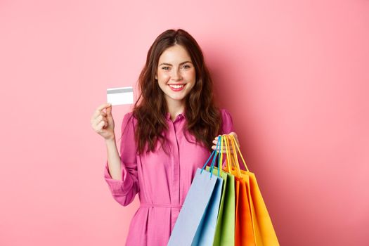 Young brunette woman holding shopping bags, showing plastic credit card and smiling, standing against pink background.