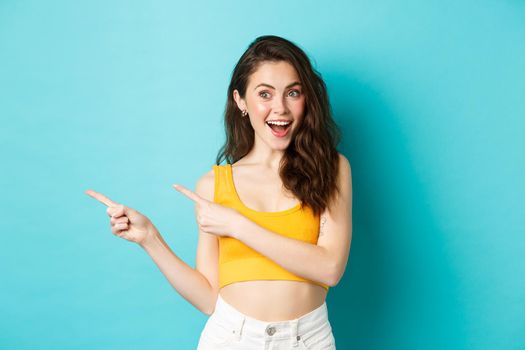 Summer lifestyle concept. Cheerful pretty girl smiling amused, pointing fingers left, recommending click banner, showing logo, standing over blue background, advertising.