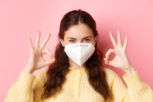 Preventive measures, health care concept. Close up of confident attractive woman, wearing medical respirator, looking determined and showing okay signs, standing against pink background.