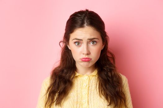 Close up of cute and coy girl making sad pouting face to ask for something, wants pity, standing against pink background gloomy.