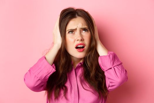 Close up of annoyed glamour girl covering ears with hands, looking bothered up at neighbours, cant stand loud music, standing over pink background.