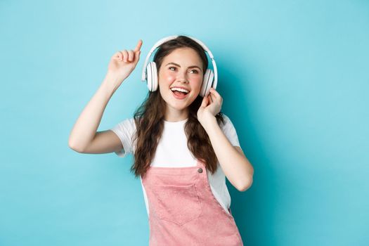 Young woman dancing and listening music in headphones, raise hand up and smiling carefree, enjoying favourite song, standing over blue background.