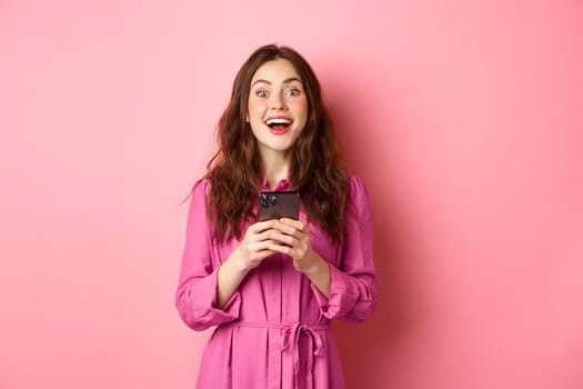 Beautiful brunette girl with amazed face, gasping and looking wondered, holding smartphone, see awesome news online, standing against pink background.