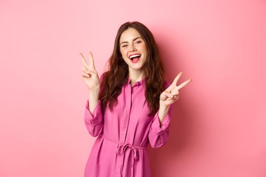 Positive beautiful girl in stylish dress, showing v-sign peace and smiling happy, posing near pink background.
