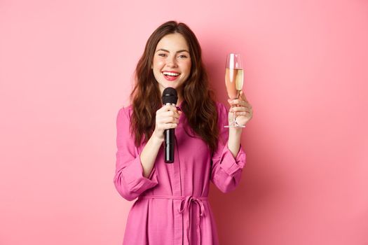 Celebration and holidays concept. Beautiful young woman saying toast, raising glass of chamapgne while making speech, holding microphone, standing over pink background. Copy space