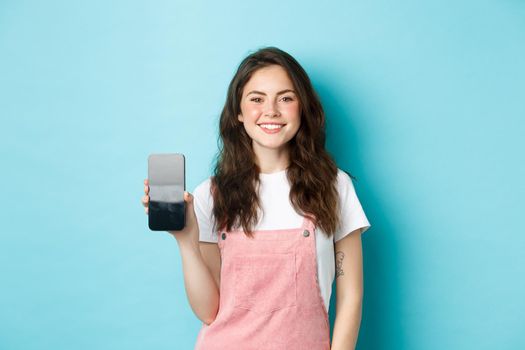 Portrait of smiling beautiful woman showing her smartphone screen and looking pleased, recommending application or online page, standing over blue background.