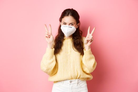 Covid-19, lockdown and pandemic concept. Cute glamour girl wears medical respirator for going outdoors during quarantine, shows peace v-sign, stands against pink background.