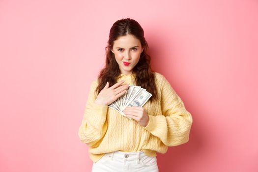 Shopping. Sassy young woman smiling and looking confident, hugging money, holding dollar bills on chest, staring thoughtful at camera, standing against pink background.