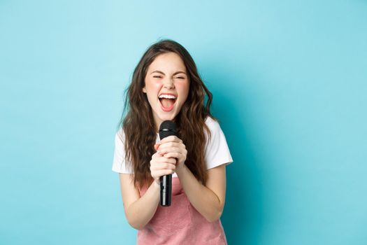 Excited pretty girl singing karaoke, holding microphone and smiling happy, standing over blue background.