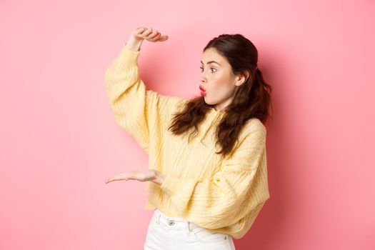 Portrait of brunette girl holds big empty space and stare amazed at place for your product or logo, showing large box gesture, standing against pink background.