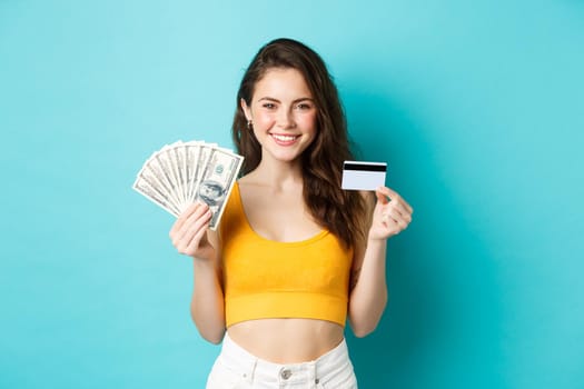 Portrait of young woman with smiling face, showing plastic credit card and money, standing against blue background.