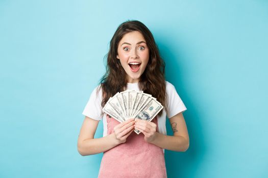 Excited cute woman winning money, holding dollar bills and smiling amazed, got fast credit, standing over blue background.