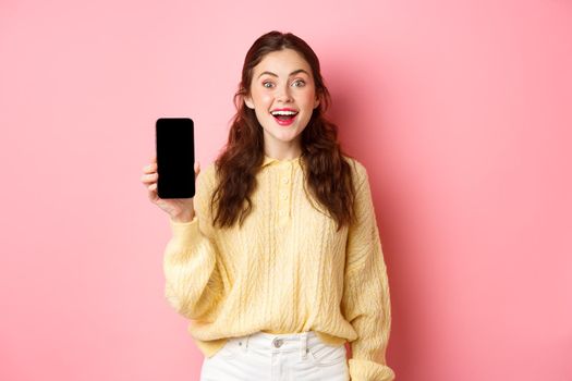 Technology and online shopping. Attractive woman student looks excited, shows empty smartphone screen and smiles, stands against pink background.