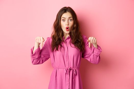 Stylish caucasian woman saying wow, pointing fingers down at impressive banner news, showing advertisement, staring amazed, standing against pink background.