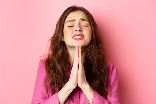 Image of hopeful young woman in need, praying, begging for help, standing over pink background in supplication pose.