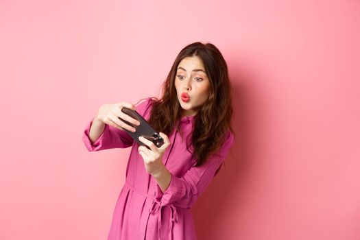 Technology concept. Carefree young woman tilt body, holding smartphone horizontally, playing video game on phone, standing against pink background.