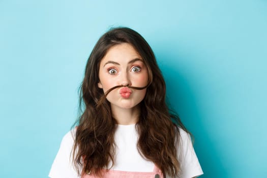 Close up portrait of funny silly girl playing around, making moustache with hair strand above lips, standing over blue background.