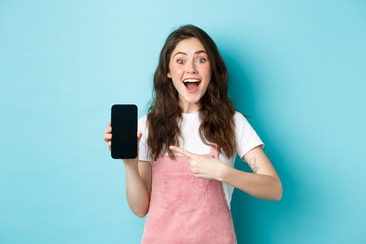 Wow check this out. Excited pretty girl pointing finger at phone screen, showing logo or store advertisement on smartphone, standing against blue background.