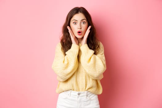 Young woman gasping and saying wow, looking surprised at camera, checking out promotion, staring at promo offer, standing against pink background.