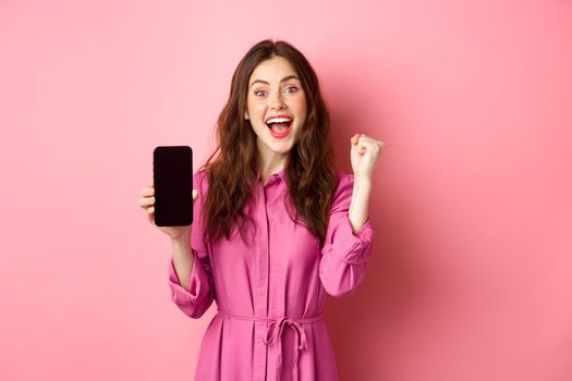 Technology concept. Excited girl shows mobile phone screen and scream with joy and happiness, winning money online, achieve daily goal on smartphone app, pink background.