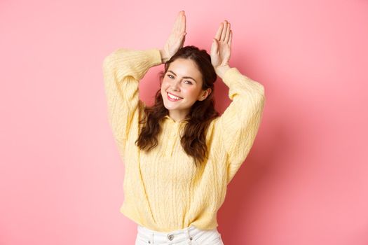 Portrait of cute lady showing Easter Bunny ears gesture and smiling, posing like silly rabbit, standing against pink background.