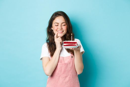 Holidays and celebration. Smiling birthday girl celebrating, looking dreamy at cake with candle, making wish, wants to bite dessert, standing over blue background.