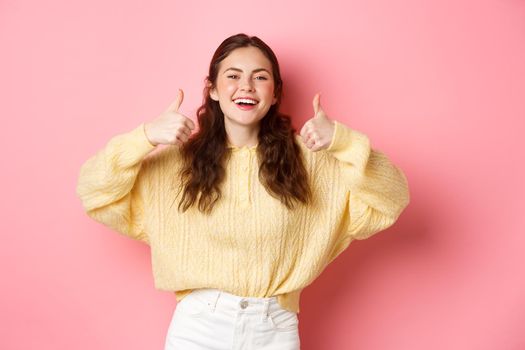 Very good, well done. Smiling girl being supportive, laughing and showing thumbs up in approval, like awesome idea, praise you, standing against pink background.