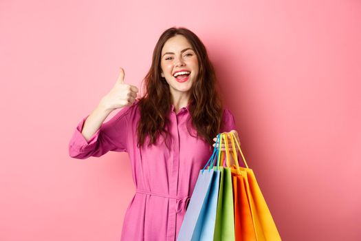 Young happy female shopper showing thumbs up, pleased with good discounts, buying staff on sale, holding shopping bags and smiling pleased, pink background.