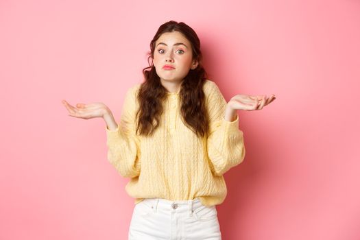 I dont know. Indecisive girl shrugging shoulders and looking unaware at camera, standing clueless against pink background. Copy space