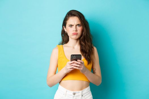 Confused and freak out young woman frowning, grimacing after reading strange message on smartphone, standing over blue background.