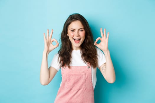 Very well, alright. Excited smiling brunette guy say yes, showing okay signs and agree with you, praising good job, standing happy against blue background.