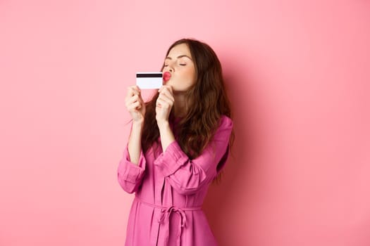 Image of young beautiful woman kissing her credit card, going on shopping, wasting money in stores, standing against pink background. Copy space