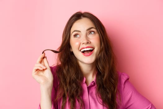 Fashion and beauty concept. Stylish young woman with make up and hairstyle, playing with hair strand and looking aside with coquettish, thoughtful smile, standing against pink background.