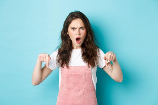 Shocked and confused young woman gasping and looking startled at camera, pointing fingers down, standing in summer clothes against blue background.