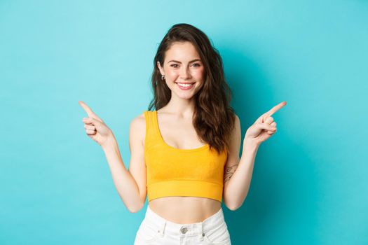 Choose your turn. Smiling beautiful woman with perfect body in crop top, pointing fingers sideways, showing two ways, standing over blue background.