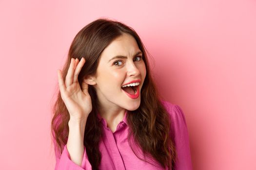 Close up of excited young woman overhear juice rumor, lean closer to camera with hand near ear, eavesdropping, standing over pink background.