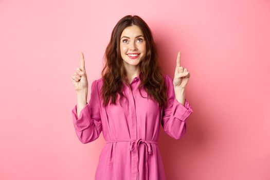 Image of beautiful happy lady in dress, pointing and looking up and smiling cheerful, staring at promo text, reading your logo, standing against pink background.