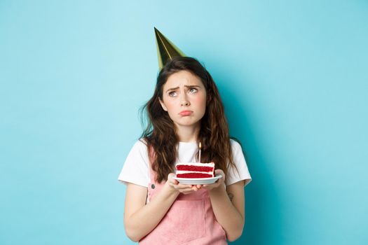 Holidays and celebration. Sad girl in party hat holding birthday cake, looking away with thoughtful upset grimace, feeling lonely and moody on her bday, standing over blue background.