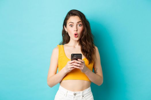 Looks interesting. Intrigued pretty girl holding smartphone, staring excited and saying wow after reading online advertisement, standing over blue background.
