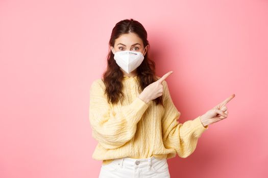 Covid, corona and social distancing concept. Excited female model in respirator shows you advertisement, points at logo aside, wears medical respirator on face, pink background.