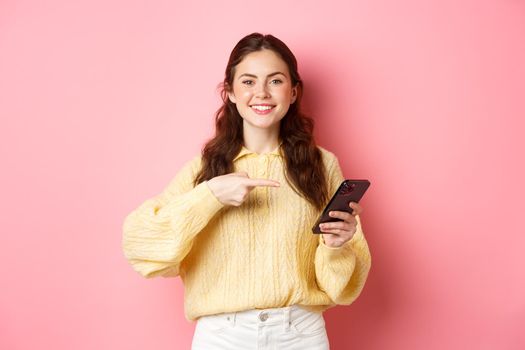 Image of happy, smiling young woman, holding mobile phone and pointing at mobile screen with pleased and confident face, standing over pink background.