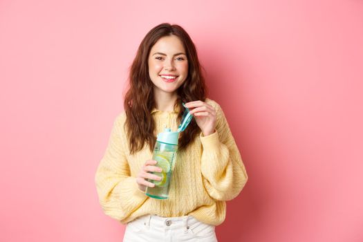 Young attractive and healthy woman smiling at camera, open water bottle with lemon, drinking sport drink, standing in colorful clothes against pink background.