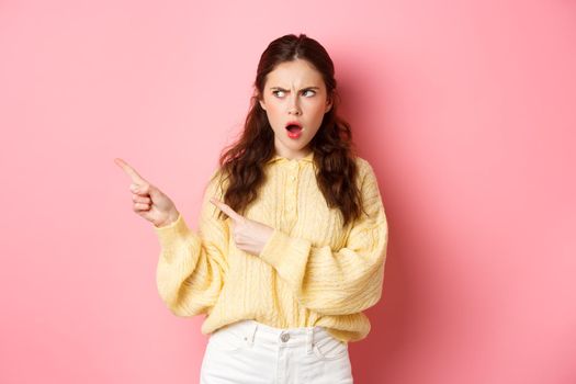 Offended and shocked young woman staring and pointing fingers left at copyspace aside, insulted by something, gasping displeased, standing against pink background.