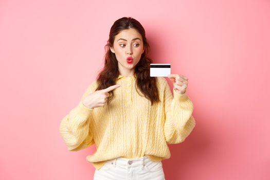 Image of glamour girl going shopping, looking excited and pointing at plastic credit card, standing against pink background.