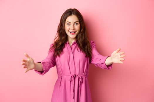 Stylish feminine lady reaching hands, stretched out arms to welcome friend, wants to hug or invite someone, standing against pink background.