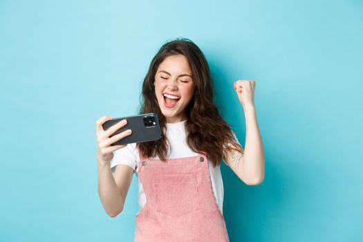 Cheerful cute girl winning in online video game on smartphone, making fist pump and shouting yes with joy, standing over blue background and triumphing.